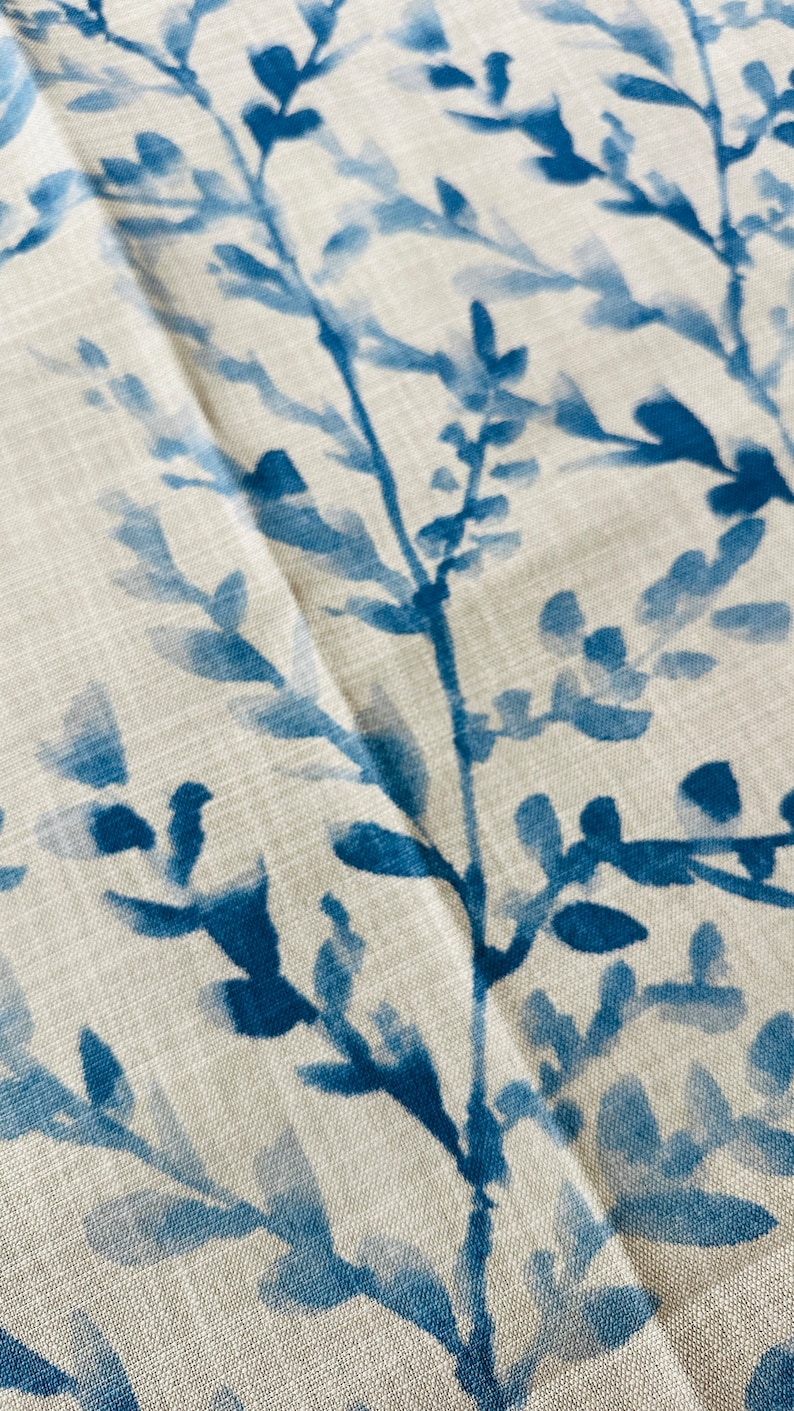 May include: Close-up of a fabric with a watercolor-style botanical print. The design features blue branches and leaves against a neutral, off-white background. The fabric has a textured appearance, suggesting a linen or cotton blend.