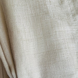 May include: Close-up of a textured, off-white fabric. The material has a subtle, woven pattern, with visible vertical and horizontal threads creating a grid-like appearance. The fabric is folded, showing depth and dimension.