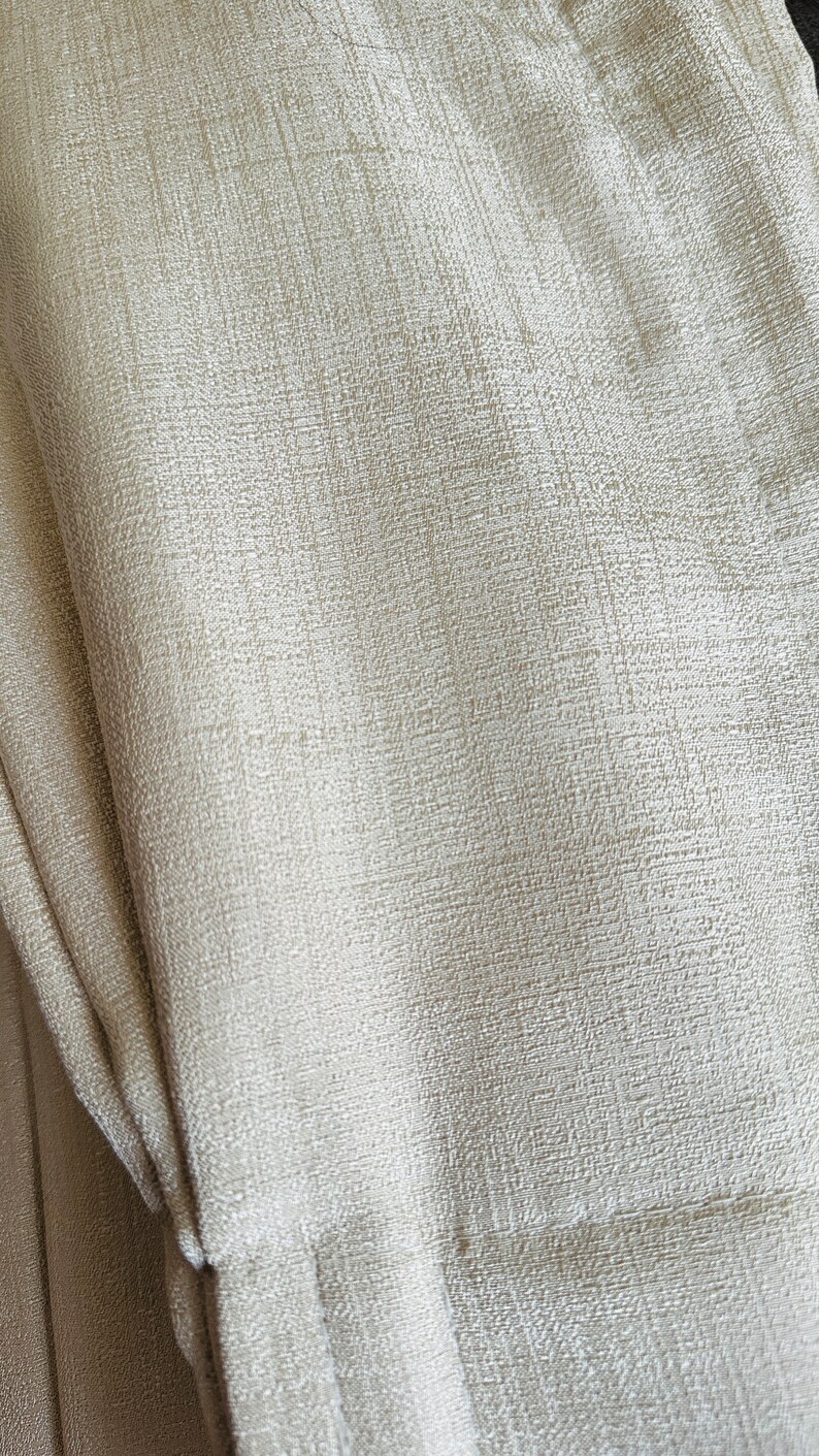 May include: Close-up of a textured, off-white fabric. The material has a subtle, woven pattern, with visible vertical and horizontal threads creating a grid-like appearance. The fabric is folded, showing depth and dimension.