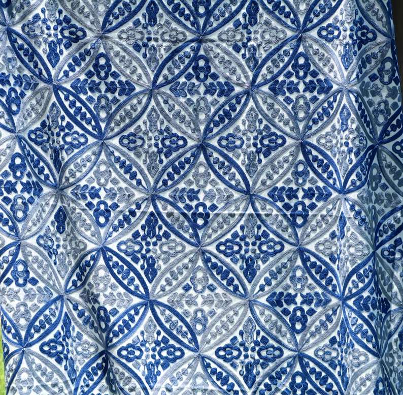 May include: White fabric with a blue and gray embroidered pattern. The pattern is a repeating geometric design with rounded shapes.