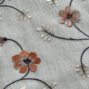 K&ouml;nnte beinhalten: Nahaufnahme eines hellgrauen Stoffes mit gestickten braunen und wei&szlig;en Blumenmustern. Der Stoff hat eine subtile Textur und die Blumen sind mit filigranen Stichen detailliert.