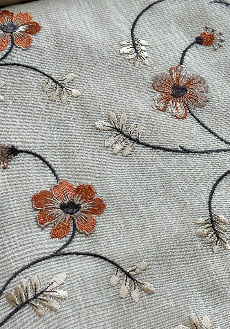 K&ouml;nnte beinhalten: Nahaufnahme eines hellgrauen Stoffes mit gestickten braunen und wei&szlig;en Blumenmustern. Der Stoff hat eine subtile Textur und die Blumen sind mit filigranen Stichen detailliert.