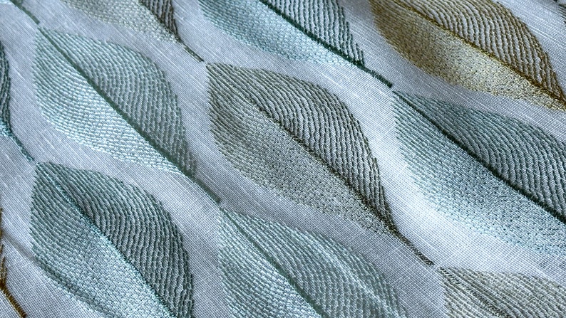 K&ouml;nnte beinhalten: Ein wei&szlig;er Stoff mit einem sich wiederholenden Muster aus blauen und goldenen Bl&auml;ttern. Der Stoff hat eine subtile Textur und einen leicht schimmernden Glanz.