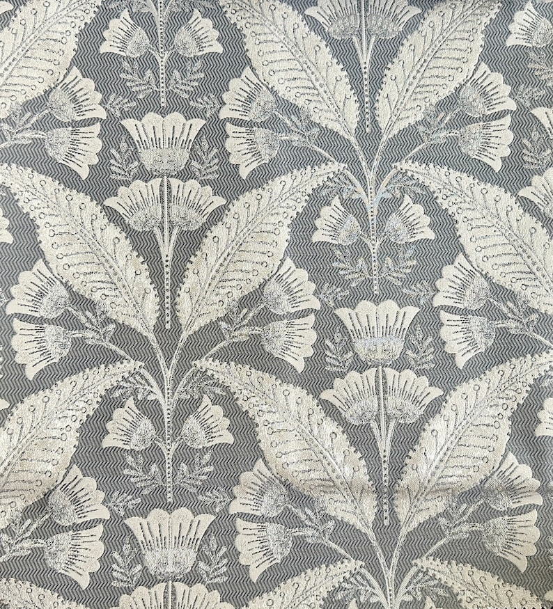 Pu&ograve; includere: Un tessuto grigio con un motivo floreale bianco. Il motivo presenta foglie e fiori stilizzati con un aspetto testurizzato, quasi metallico.