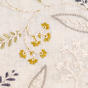 May include: Close-up of a white fabric with embroidered floral designs in gold, white, and gray. The embroidery features delicate leaves and branches.
