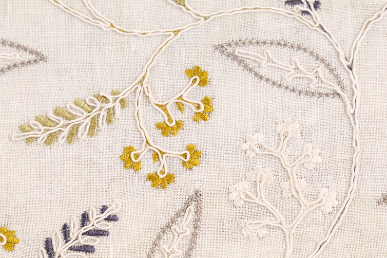 May include: Close-up of a white fabric with embroidered floral designs in gold, white, and gray. The embroidery features delicate leaves and branches.