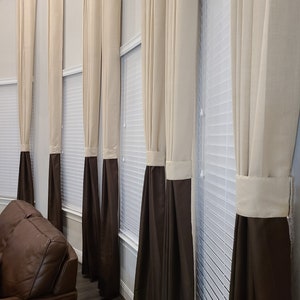 May include: A series of beige and brown curtains hang in front of a window with white blinds. The curtains are a two-tone design, with a beige upper section and a brown lower section. The curtains are hanging in a living room.