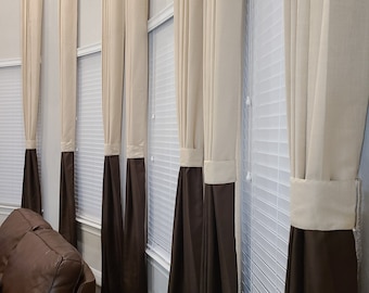 May include: A series of beige and brown curtains hang in front of a window with white blinds. The curtains are a two-tone design, with a beige upper section and a brown lower section. The curtains are hanging in a living room.