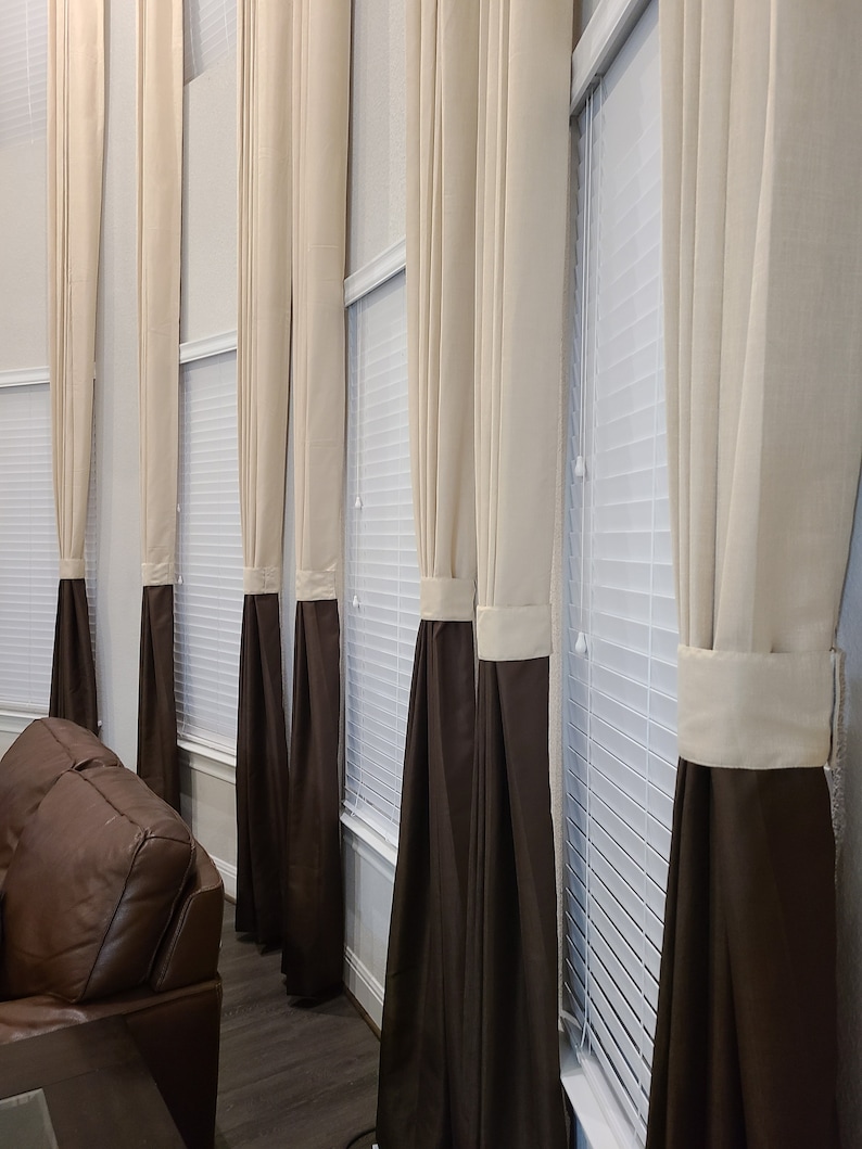 May include: A series of beige and brown curtains hang in front of a window with white blinds. The curtains are a two-tone design, with a beige upper section and a brown lower section. The curtains are hanging in a living room.
