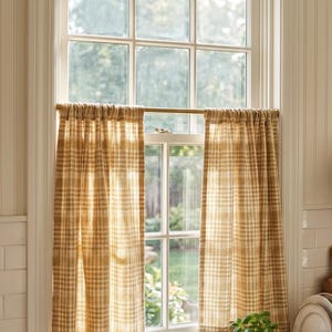 May include: A pair of cafe curtains featuring a tan and white plaid pattern, hung on a thin brass rod across the middle of a window. The fabric is semi-sheer, allowing light to filter through the checkered weave into the room.