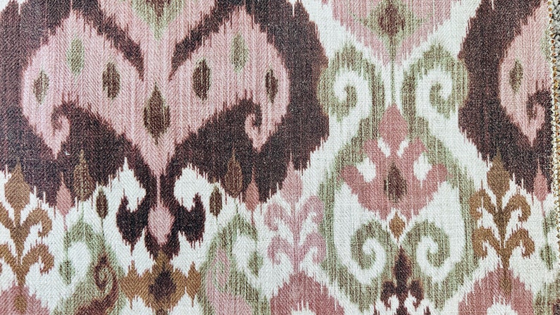May include: Close-up of a fabric with an ikat pattern. The design features repeating abstract shapes in shades of brown, pink, and green on a white background. The fabric has a textured appearance, suitable for upholstery or home decor.