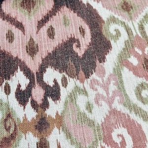 May include: Close-up of a textile with an ikat pattern. The design features swirling and geometric shapes in shades of brown, pink, green, and cream. The fabric has a textured appearance.