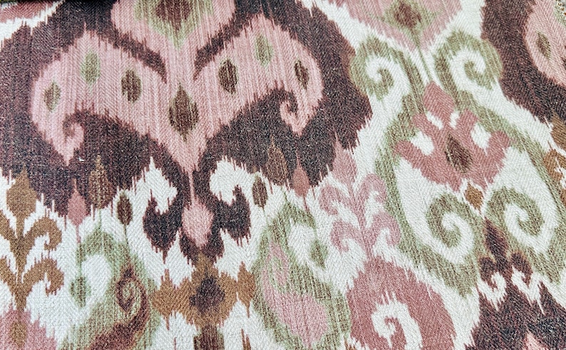 May include: Close-up of a textile with an ikat pattern. The design features swirling and geometric shapes in shades of brown, pink, green, and cream. The fabric has a textured appearance.