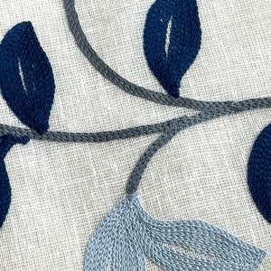 May include: A close-up of a white fabric with blue and gray embroidered leaves. The leaves are stitched in a detailed pattern, creating a delicate and elegant design.