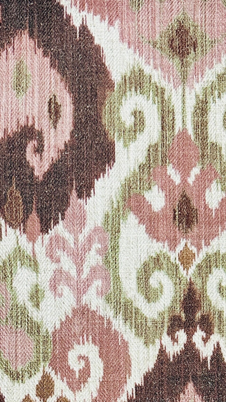 May include: Close-up of a woven fabric with an ikat design. The pattern features repeating shapes in shades of brown, pink, green, and white. The fabric has a textured appearance, suggesting a natural fibre.