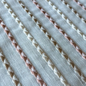 Puede incluir: Primer plano de una tela blanca con filas diagonales de bordado trenzado beige y rosa.