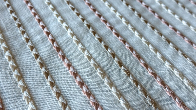 Puede incluir: Primer plano de una tela blanca con filas diagonales de bordado trenzado beige y rosa.