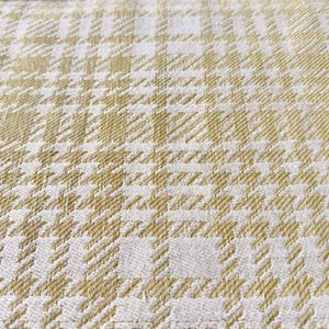 May include: A woven fabric featuring a repeating houndstooth and plaid pattern in shades of soft gold and off-white. The textile displays a textured surface with a dense, interlaced weave structure that creates a geometric grid design throughout.