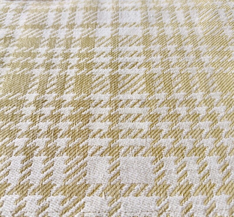 May include: A woven fabric featuring a repeating houndstooth and plaid pattern in shades of soft gold and off-white. The textile displays a textured surface with a dense, interlaced weave structure that creates a geometric grid design throughout.
