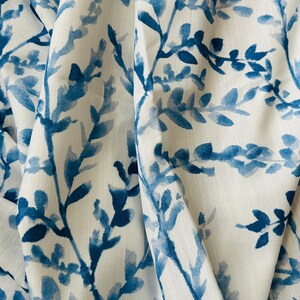 May include: White fabric with a blue watercolor-style botanical pattern. The design features branches with leaves in varying shades of blue, creating a soft, artistic look. The fabric is draped, showing folds and texture.