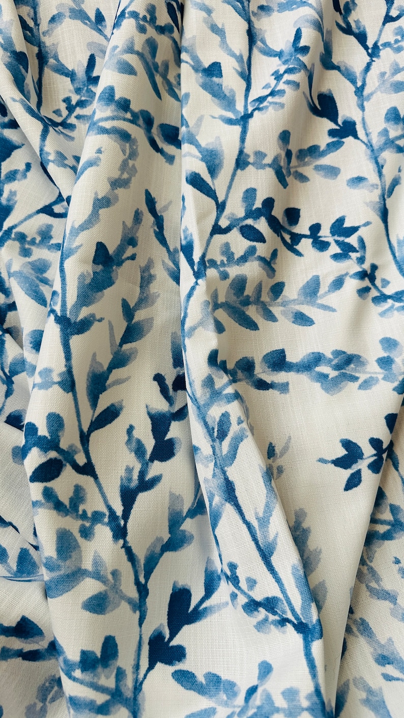 May include: White fabric with a blue watercolor-style botanical pattern. The design features branches with leaves in varying shades of blue, creating a soft, artistic look. The fabric is draped, showing folds and texture.