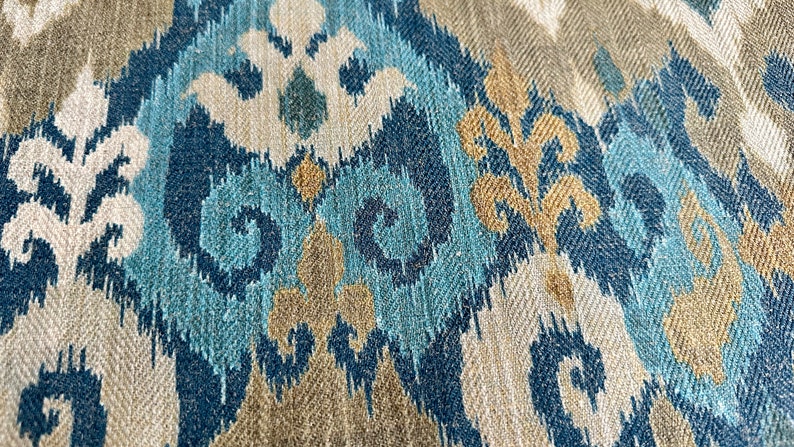 May include: Close-up of a fabric with a blue, brown, and white ikat pattern. The fabric has a woven texture and a subtle herringbone pattern.