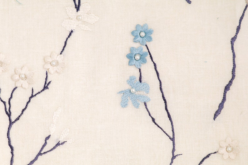 May include: White fabric with embroidered blue and white flowers. The flowers are arranged on dark blue stems.