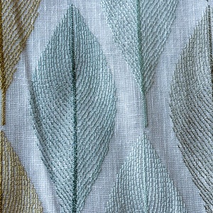 K&ouml;nnte beinhalten: Ein wei&szlig;er Stoff mit einem gestickten Muster aus blauen und goldenen Bl&auml;ttern. Der Stoff hat eine gewebte Textur.