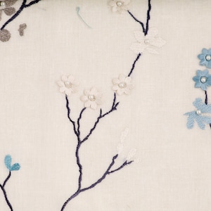 May include: A white fabric with a delicate floral embroidery design. The flowers are in shades of blue and white, with small pearl-like accents. The fabric features a subtle texture and a light, airy feel.