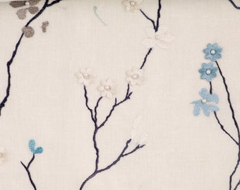 May include: A white fabric with a delicate floral embroidery design. The flowers are in shades of blue and white, with small pearl-like accents. The fabric features a subtle texture and a light, airy feel.