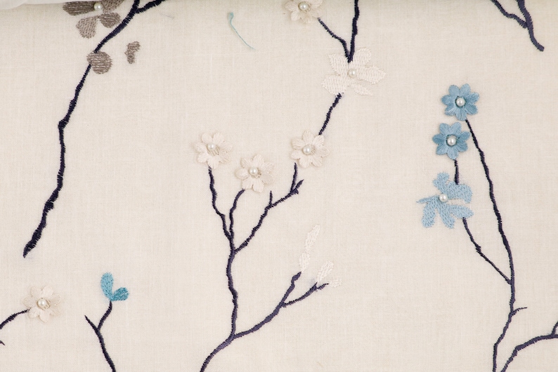 May include: A white fabric with a delicate floral embroidery design. The flowers are in shades of blue and white, with small pearl-like accents. The fabric features a subtle texture and a light, airy feel.
