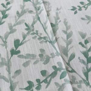 May include: White fabric with a green leafy pattern. The fabric has a subtle texture and the leaves are a light green color.
