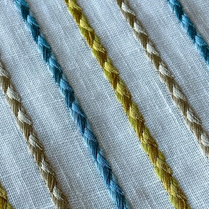 May include: Close-up of a white fabric with diagonal stripes of blue, yellow, and beige braided trim.