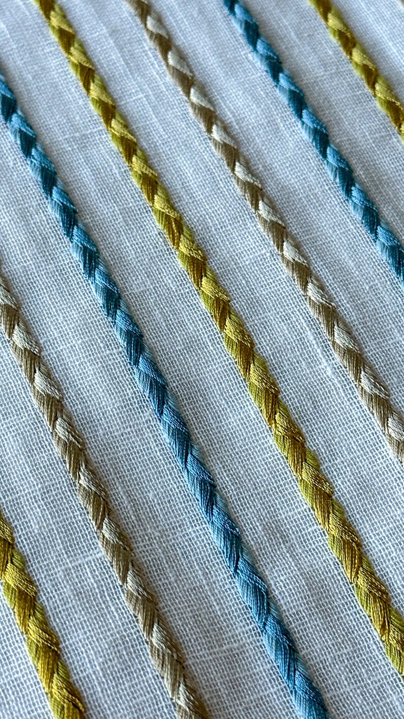 May include: Close-up of a white fabric with diagonal stripes of blue, yellow, and beige braided trim.