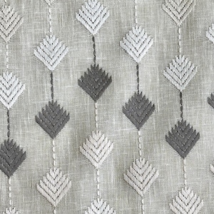 May include: Close-up of a textured fabric with a repeating pattern of diamond shapes. The diamonds are embroidered in shades of white and gray, connected by vertical lines. The background is a light beige color.