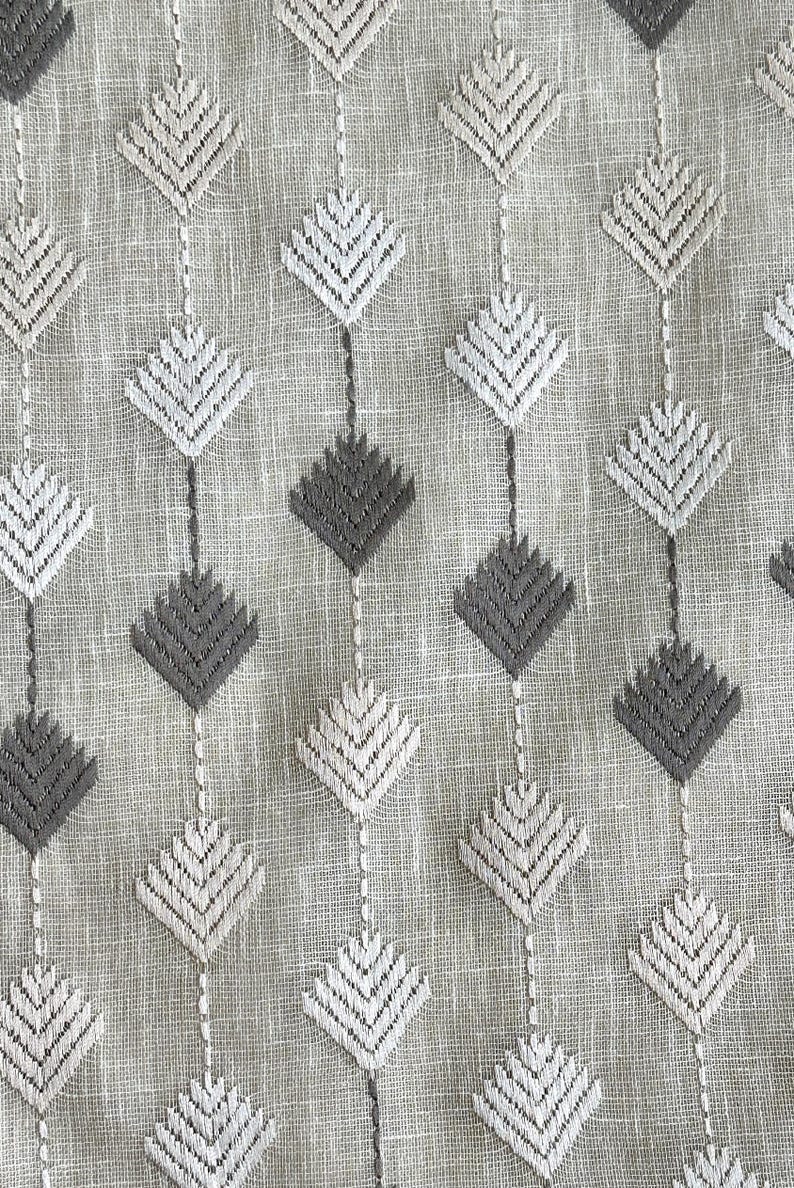 May include: Close-up of a textured fabric with a repeating pattern of diamond shapes. The diamonds are embroidered in shades of white and gray, connected by vertical lines. The background is a light beige color.