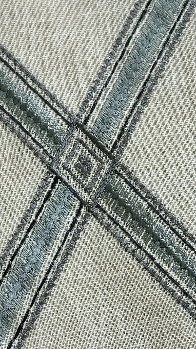K&ouml;nnte beinhalten: Nahaufnahme eines hellgrauen Stoffes mit zwei sich kreuzenden B&auml;ndern aus dunkelgrauer und hellgrauer Stickerei. Die Stickerei ist ein sich wiederholendes Muster aus kleinen Dreiecken und Quadraten.
