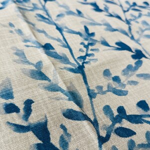 May include: Close-up of a fabric with a botanical design. The fabric features a light beige background with a pattern of blue branches and leaves. The leaves and branches are painted in varying shades of blue.