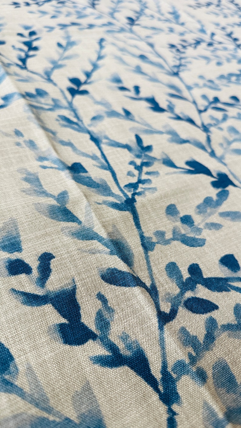 May include: Close-up of a fabric with a botanical design. The fabric features a light beige background with a pattern of blue branches and leaves. The leaves and branches are painted in varying shades of blue.