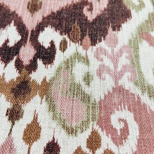 May include: Close-up of a textile with an ikat pattern. The design features repeating shapes in shades of brown, pink, and green on a white background. The fabric has a textured appearance.