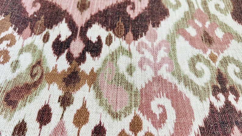 May include: Close-up of a textile with an ikat pattern. The design features repeating shapes in shades of brown, pink, and green on a white background. The fabric has a textured appearance.