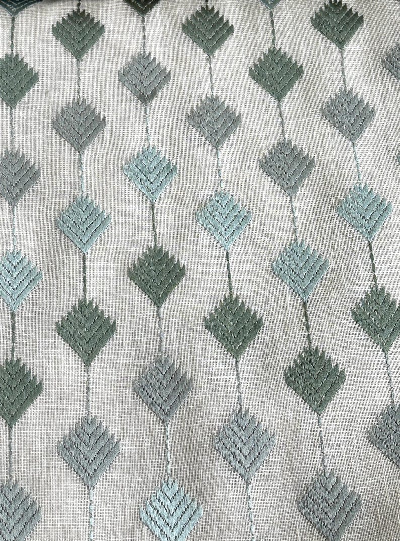 May include: White fabric with a repeating pattern of blue and green embroidered diamond shapes. The diamonds are made up of a series of small, overlapping triangles.
