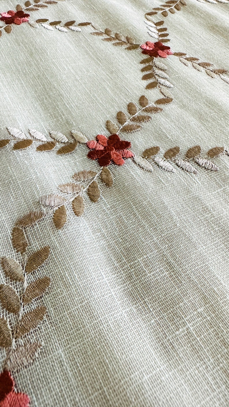 Puede incluir: Un mantel de tela blanco con un delicado patr&oacute;n bordado de hojas marrones y flores rojas.
