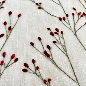K&ouml;nnte beinhalten: Wei&szlig;er Stoff mit gestickten roten Beeren und gr&uuml;nen Stielen. Der Stoff hat eine subtile Textur.