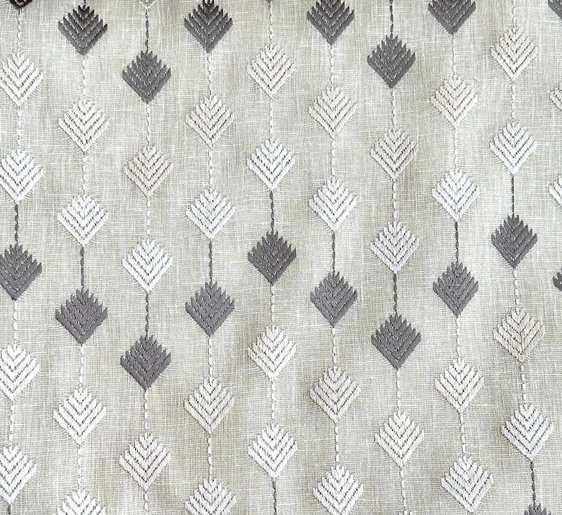 May include: Close-up of a fabric with a repeating diamond pattern. The diamonds are embroidered in alternating shades of gray and white, connected by vertical lines. The background is a light beige color, giving the fabric a textured appearance.