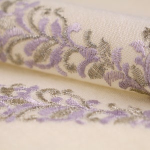 May include: Close-up of a white fabric with a delicate embroidered design featuring a repeating pattern of lavender and silver floral motifs.