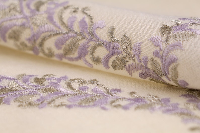 May include: Close-up of a white fabric with a delicate embroidered design featuring a repeating pattern of lavender and silver floral motifs.