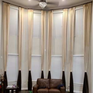 May include: A living room with tall, beige curtains framing large windows. The curtains have a dark brown accent at the bottom. A brown leather sofa sits below the windows. A ceiling fan and recessed lighting are visible.