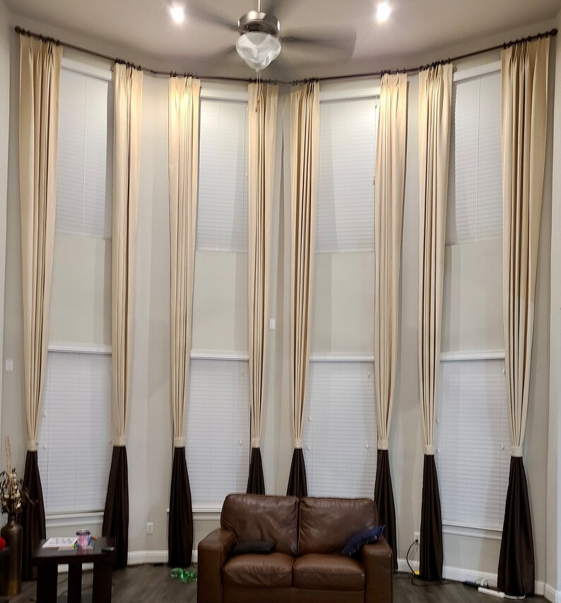 May include: A living room with tall, beige curtains framing large windows. The curtains have a dark brown accent at the bottom. A brown leather sofa sits below the windows. A ceiling fan and recessed lighting are visible.