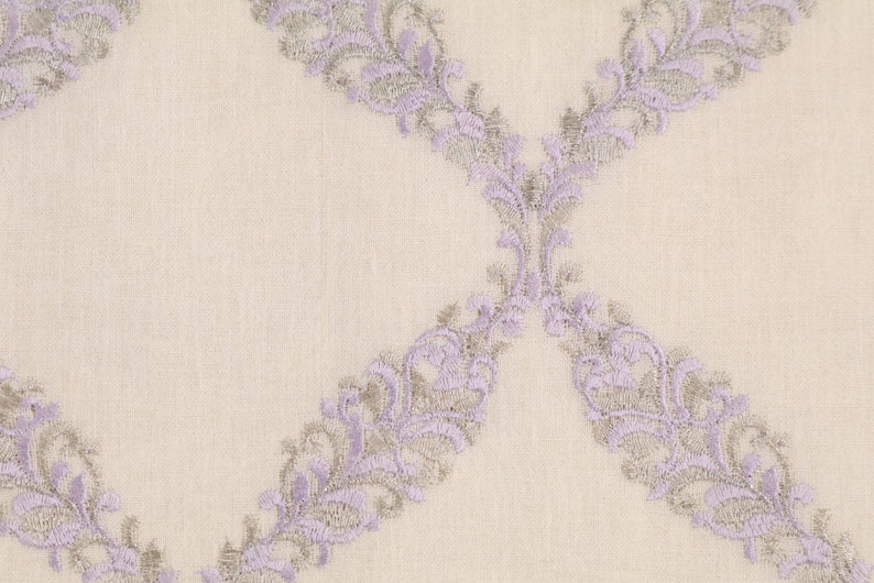 May include: A close-up of a white fabric with a delicate lavender and silver embroidered pattern. The pattern features a repeating design of stylized floral motifs.
